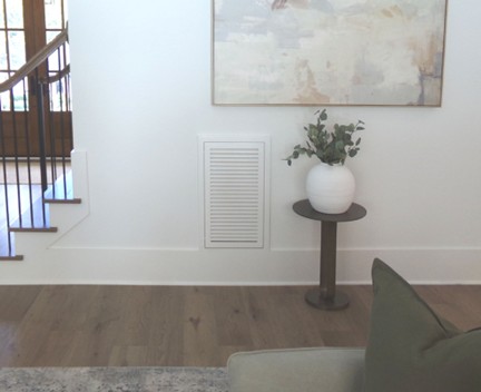 Wood Return Air Filter Grille painted and installed in wall beside stairs. Abstract color picture hangs on the wall above the grille and to the right is a vase with a plant on a small round wood table on hardwood floors